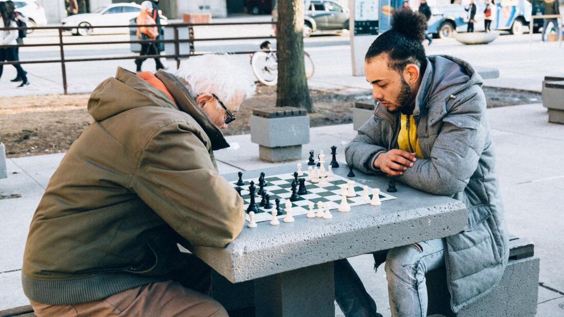 An older man and a younger Latino man play chess in a park.