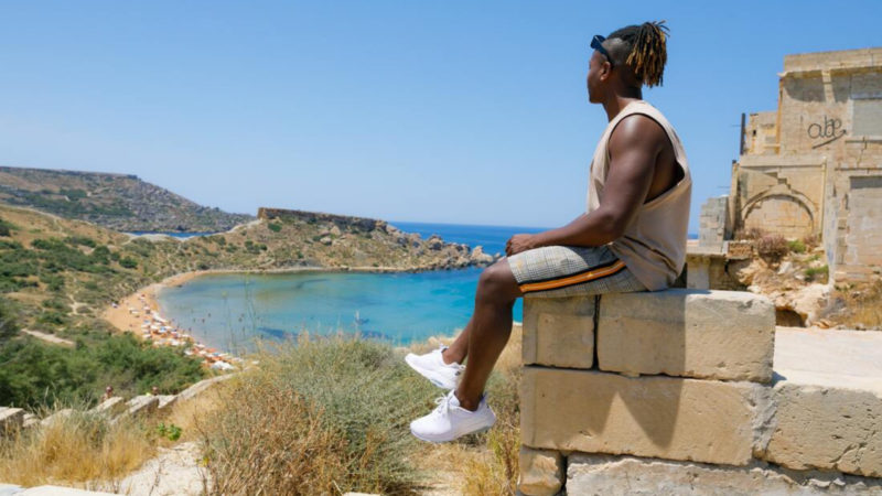 A young black tour guide sits on a stone wall overlooking the sea
